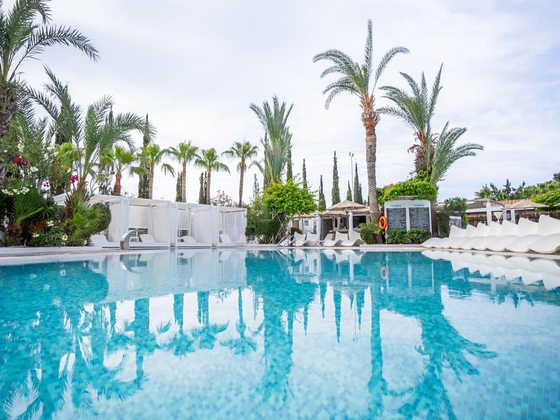 Großer Pool mit klarem Wasser, umgeben von Palmen und weißen Liegestühlen unter blauem Himmel.