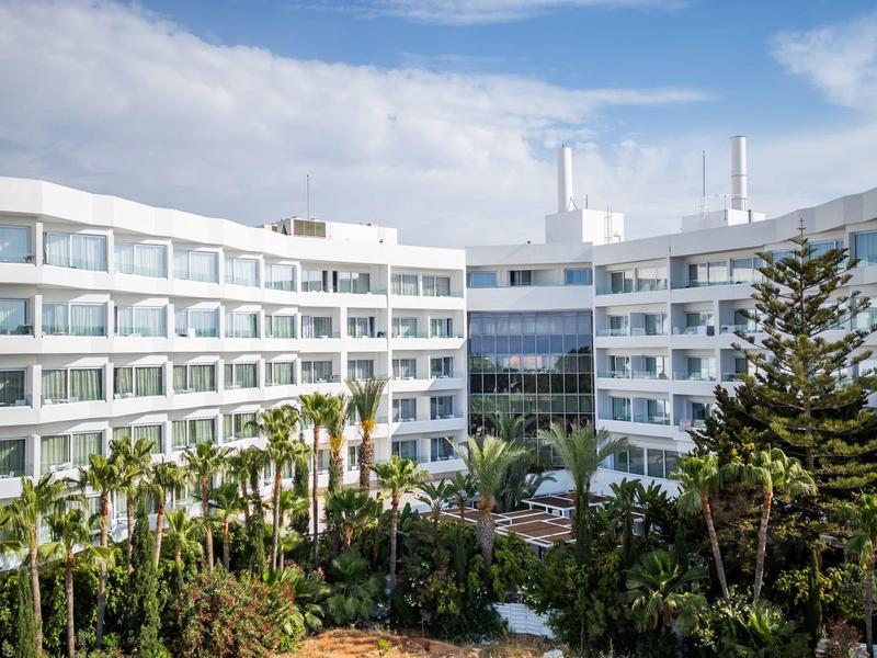 Weißes Hotelgebäude mit großen Fenstern, mehreren Stockwerken und grüner Gartenanlage davor.