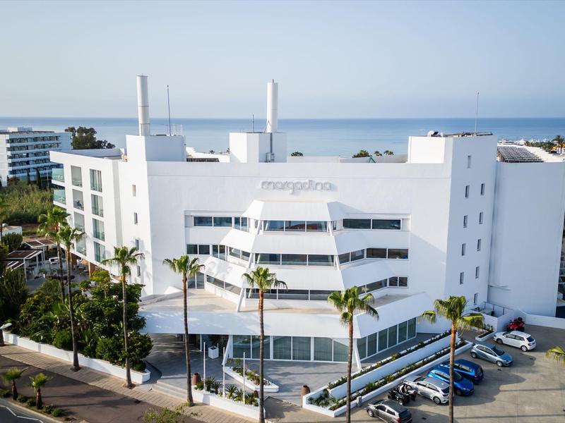 Weißes modernes Gebäude mit Glasschiebetüren, Palmen und Autos vor blauem Himmel und Meer.