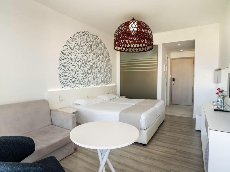 Helles Hotelzimmer mit Doppelbett, Sofaecke, rundem Tisch und moderner Beleuchtung.