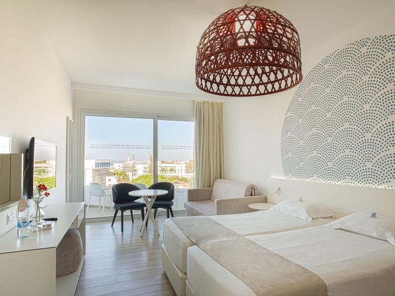 Helles Schlafzimmer mit zwei Betten, Sofa, TV, großem Fenster und modernem Holzlampe über dem Bett.