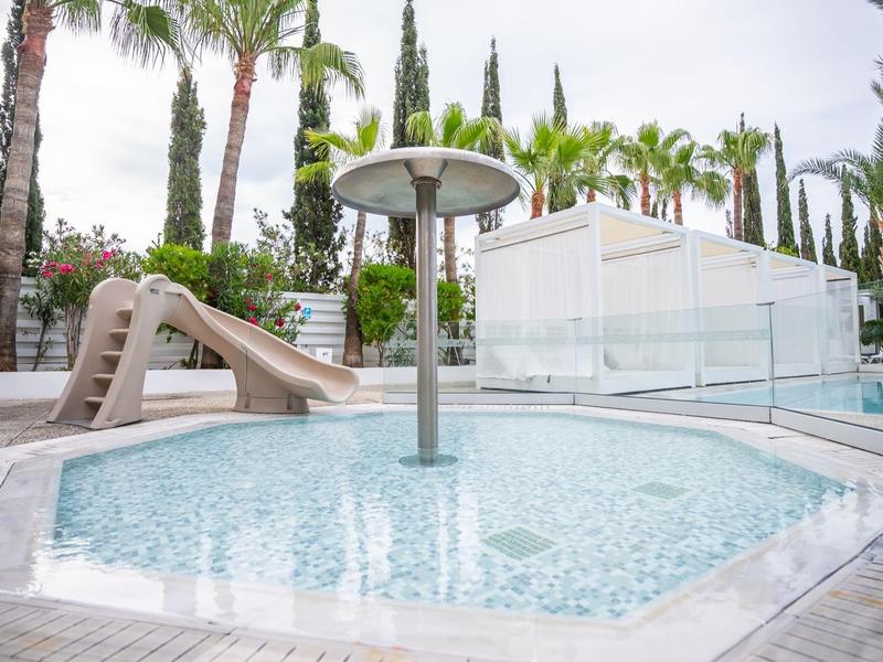 Kleines, rundes Kinderplanschbecken mit Rutsche, Wasserpilz und Palmen im Hintergrund.