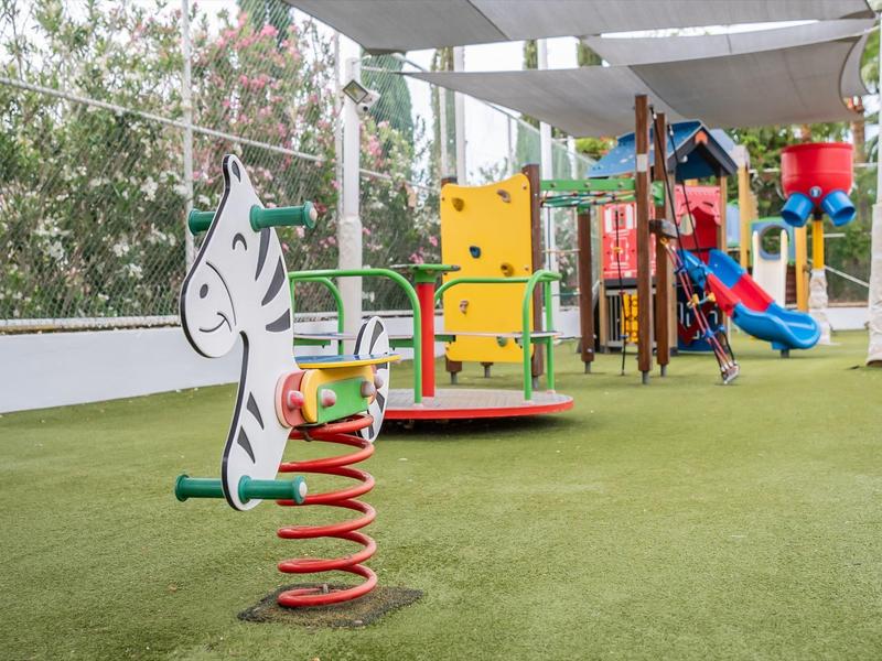 Bunter Spielplatz mit Feder-Schaukel, Klettergerüst und Rutsche auf grünem Kunstrasen.