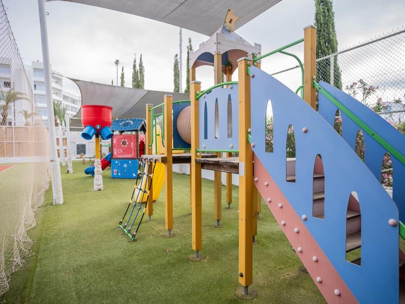 Bunter Kinderspielplatz mit Kletternetz, Rutschen und Trampolin unter Dach auf grünem Boden.