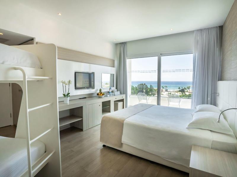Helles Schlafzimmer mit Doppelbett, Etagenbett, Schreibtisch, TV und Balkon mit Meerblick.