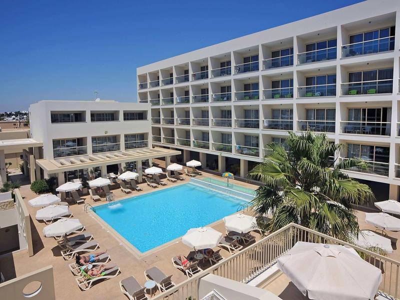 Großes Hotel mit rechteckigem Pool, Sonnenschirmen und Liegestühlen unter klarem Himmel.