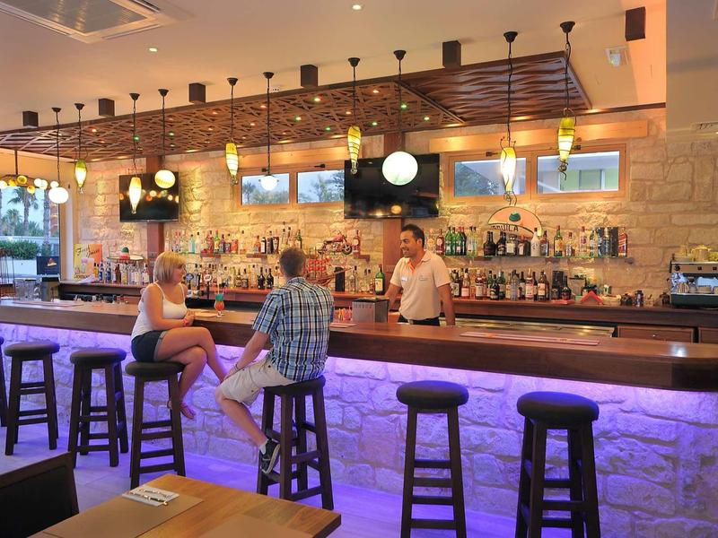 Moderne Bar mit beleuchtetem Tresen, Holzstühlen, Steinwand und drei Gästen am Tresen.