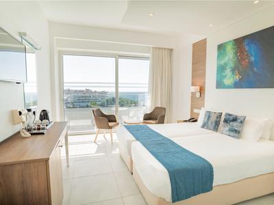 Helles Hotelzimmer mit Doppelbett, Balkon mit Meerblick und moderner Einrichtung.