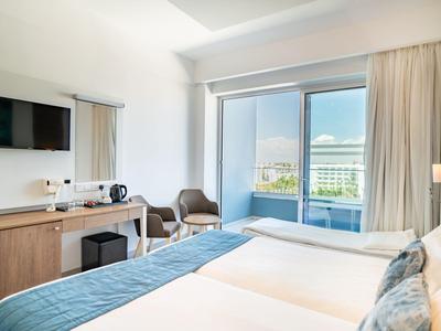 Helles Hotelzimmer mit zwei Betten, Fernseher, Schreibtisch und Balkon mit Blick auf Stadt und Berge.