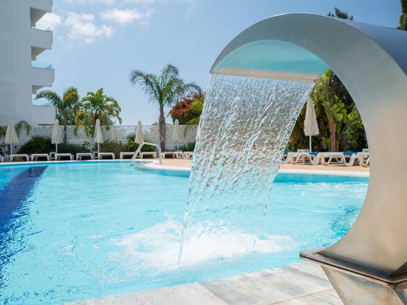 Moderner Wasserfall im Pool eines Hotels mit Palmen und Liegestühlen im Hintergrund.