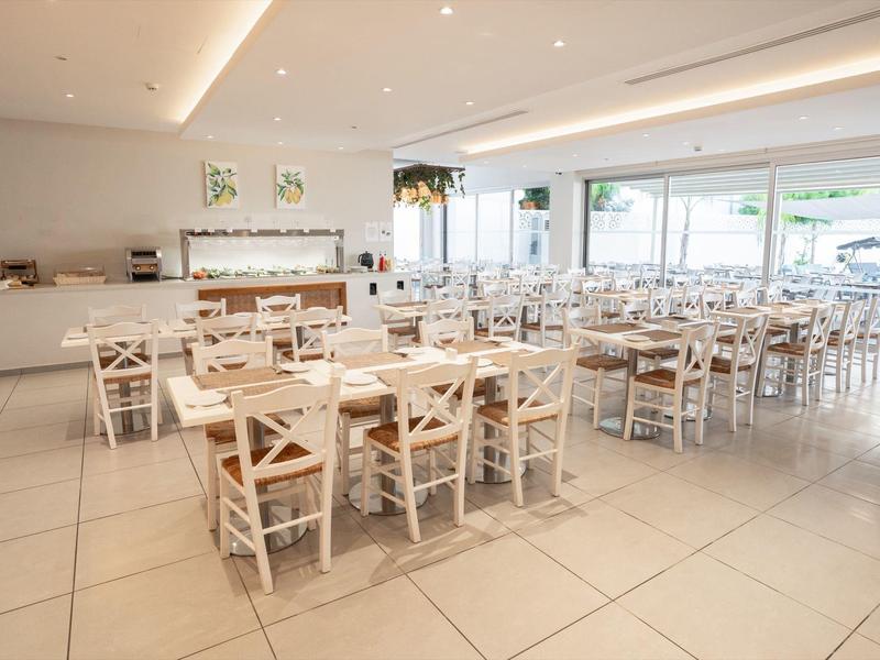 Helles, modernes Restaurant mit Holztischen und weißen Stühlen, großen Fenstern und Fliesenboden.