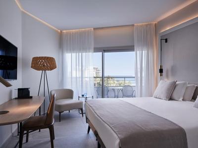 Modern hotel room with double bed, desk, armchair, and balcony with sea view.