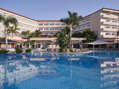 Großes Hotel mit mehreren Stockwerken, Palmen und einem großen Swimmingpool in der Sonne.