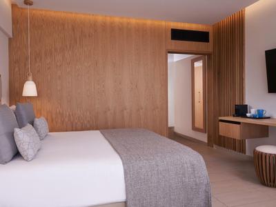 Modern hotel room with large bed, wood wall, TV, and seating area in warm lighting.