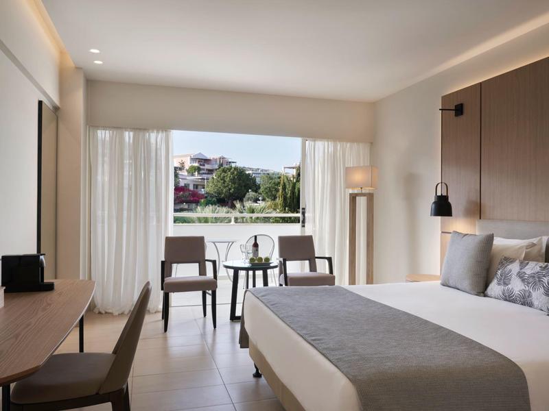 Bright hotel room with large bed, desk, chairs, and balcony overlooking garden and buildings.