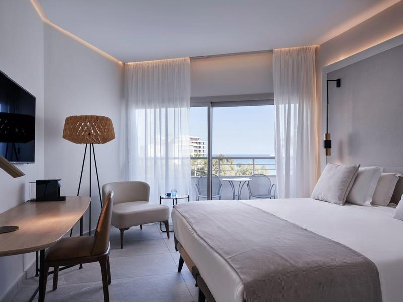 Modern hotel room with double bed, desk, armchair, and balcony with sea view.