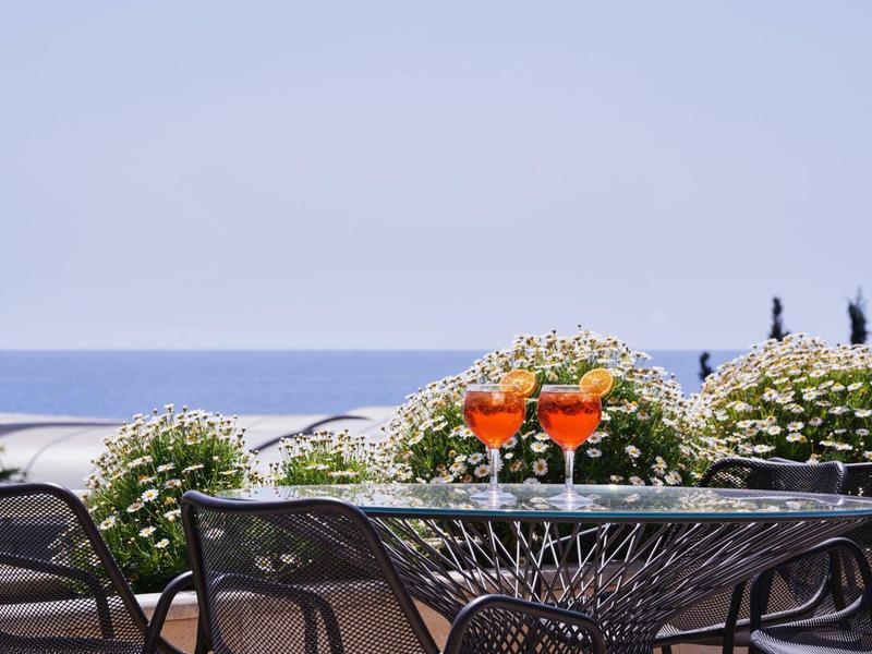 Terrace with table and chairs, two orange cocktails, view of the sea and flowering bushes.