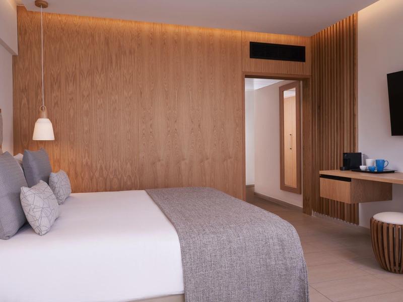 Modern hotel room with large bed, wood wall, TV, and seating area in warm lighting.