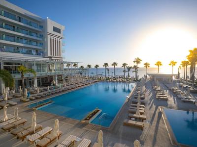 Hotel mit großem Pool, Liegestühlen und Palmen am Strand bei Sonnenuntergang.