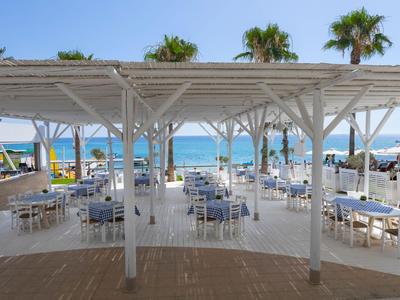 Restaurante de playa abierto con mesas y sillas blancas bajo una pérgola de madera blanca con vistas al mar.