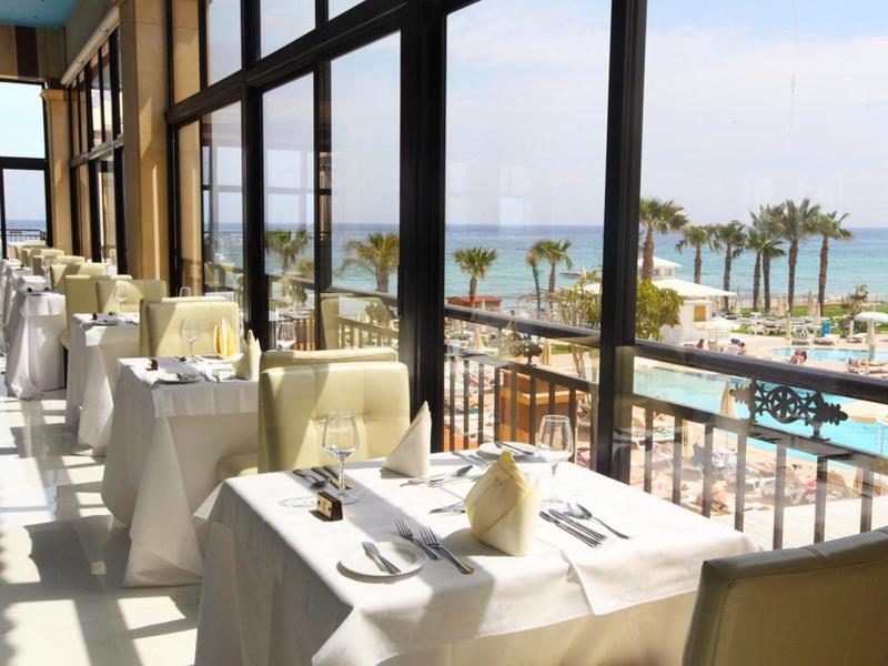 Heller Restaurantraum mit weiß gedeckten Tischen, großen Fenstern und Meerblick mit Palmen.