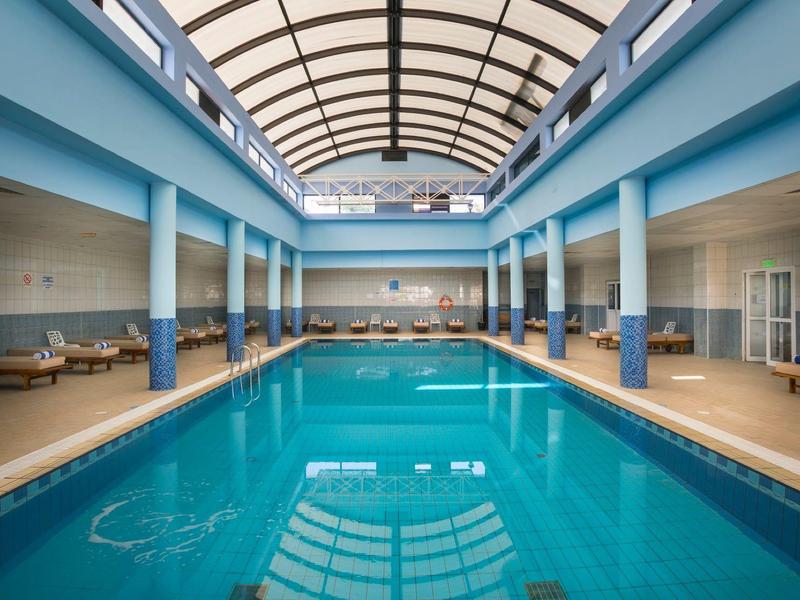 Hallenbad mit klarem blauem Wasser, beigen Fliesen und hohen blauen Säulen unter Glasdach.