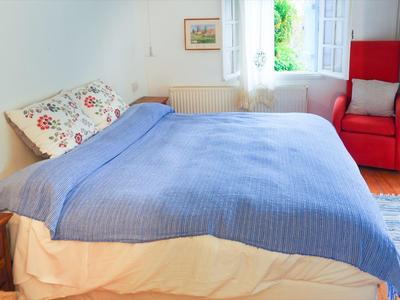 Ein gemütliches Schlafzimmer mit großem Bett, blauem Überwurf und rotem Sessel neben einem Fenster.