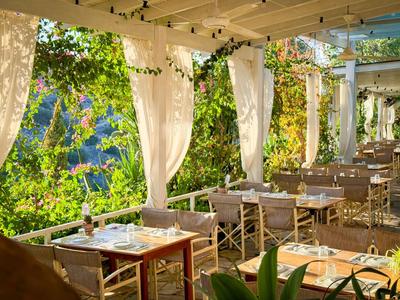 Terraza de restaurante con mesas y sillas rodeada de plantas verdes y cortinas blancas.