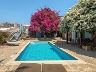 Piscina con agua clara entre dos árboles en flor junto a una casa bajo un cielo azul.