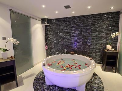 Rundes Whirlpool-Bad mit Rosenblättern vor dunkler Steinwand, dekoriert mit Orchideen und sanfter Beleuchtung.