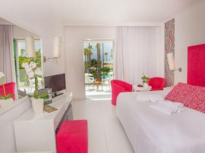 Helles Hotelzimmer mit weißem Mobiliar, rotem Kopfteil und Stuhl, sowie Balkon mit Meerblick.