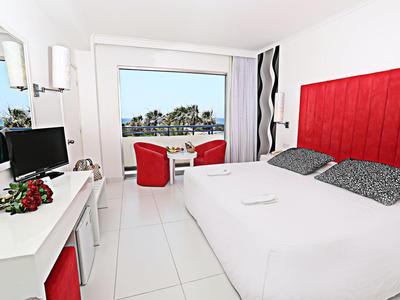 Helles Hotelzimmer mit großem Fenster, rotem Kopfteil, weißem Bett und zwei roten Sesseln.