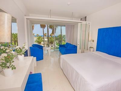 Helles Schlafzimmer mit großem Bett, blauen Akzenten und Blick auf Palmen durch Glas-Schiebetür.