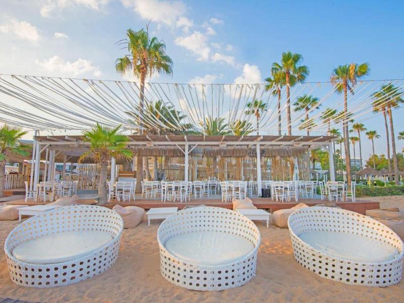 Strand mit palmengesäumtem Lounge-Bereich, weißen Korbsesseln und Sonnensegeln am Himmel.