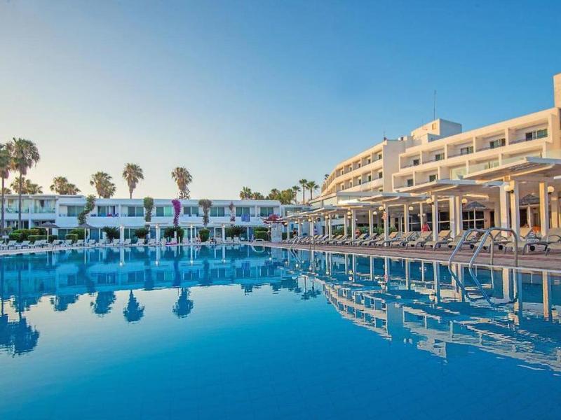 Großes Hotel mit weißer Fassade neben klarem, blauem Pool und Palmen unter blauem Himmel.