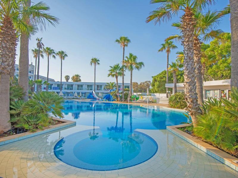 Großer blauer Pool mit Palmen, umgeben von grünen Pflanzen und weißen Hotelgebäuden.