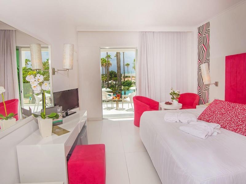 Helles Hotelzimmer mit weißem Mobiliar, rotem Kopfteil und Stuhl, sowie Balkon mit Meerblick.