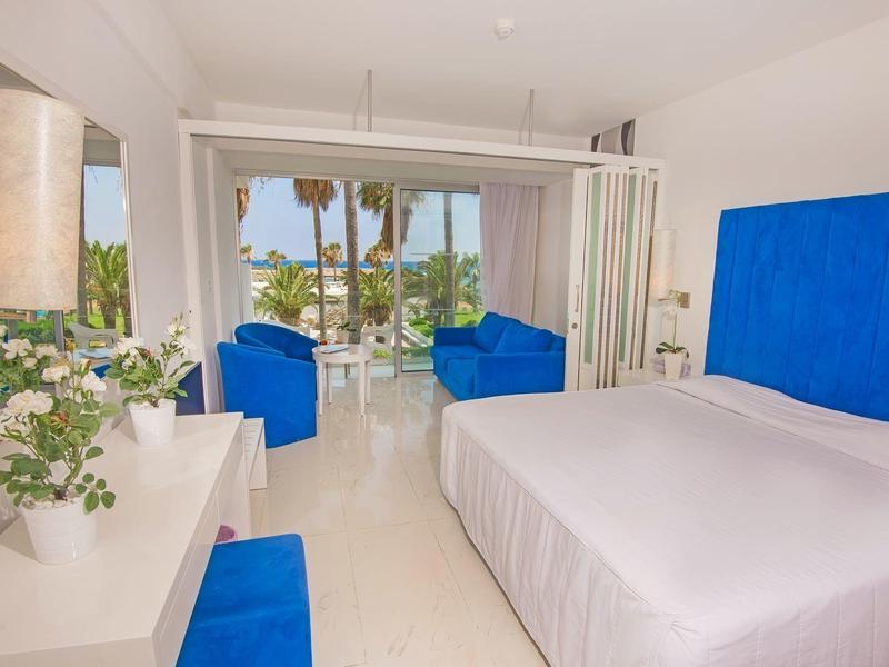 Helles Schlafzimmer mit großem Bett, blauen Akzenten und Blick auf Palmen durch Glas-Schiebetür.