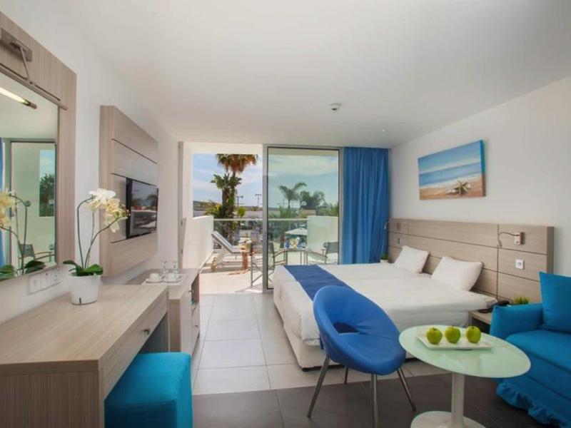 Helles Hotelzimmer mit Doppelbett, blauem Sessel, Sofa, Tisch und Balkon mit Meerblick.