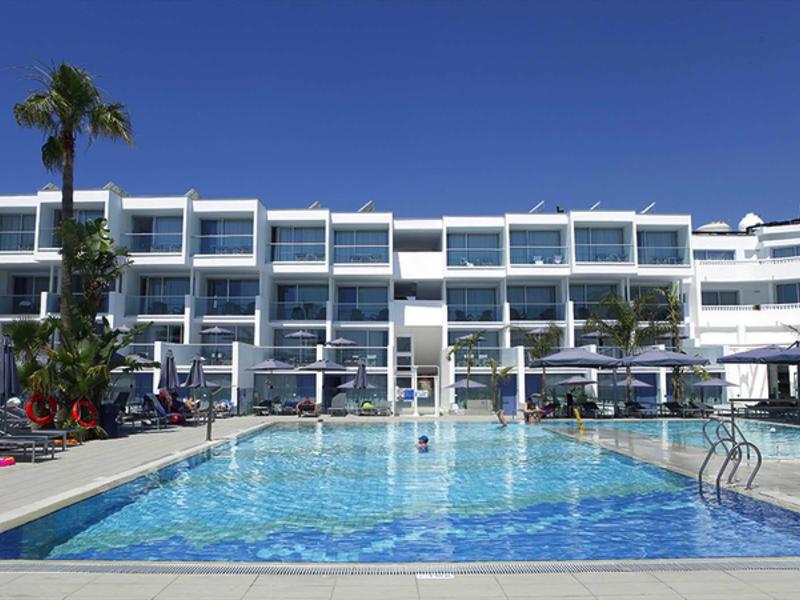 Moderner Hotelkomplex mit großem Außenpool, Palmen und klarem blauem Himmel im Hintergrund.