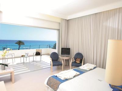 Helles Hotelzimmer mit Doppelbett, Sitzbereich und Balkon mit Meerblick.