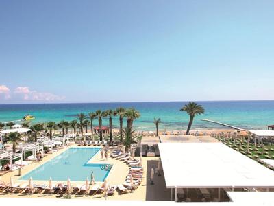 Strandhotel mit Pool, Palmen und Blick auf das ruhige, blaue Meer unter klarem Himmel.