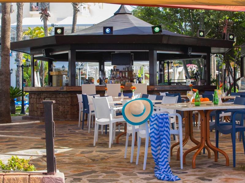 Freiluftrestaurant mit bunten Stühlen, runden Tischen und Sonnenschirm in warmem Tageslicht.