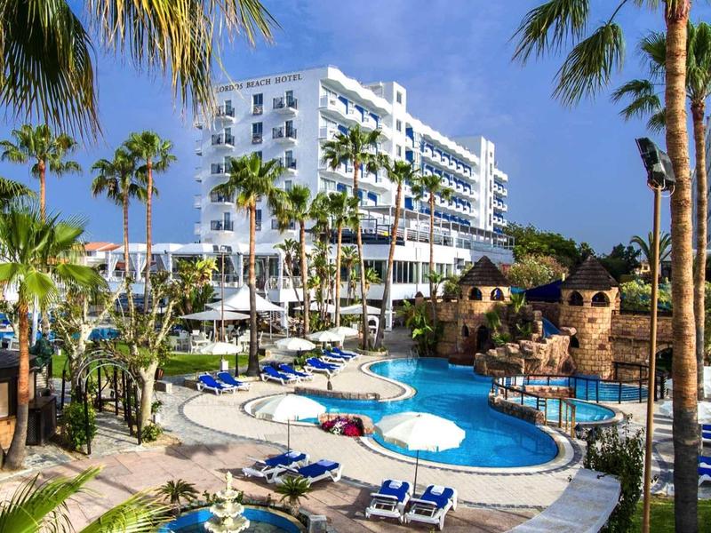 Hotelanlage mit blauem Pool, Liegestühlen, Palmen und mehrstöckigem weißem Gebäude im Hintergrund.