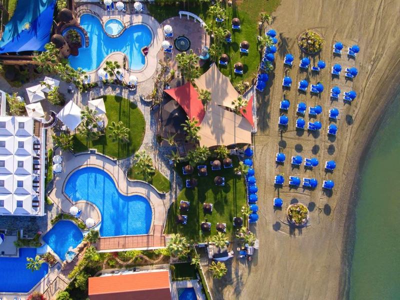 Luftaufnahme eines Resorts mit blauen Pools, Liegestühlen und Sandstrand am türkisfarbenen Meer.
