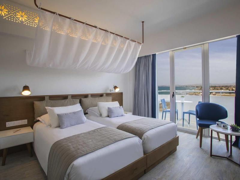 Modernes Hotelzimmer mit zwei Einzelbetten, Meerblick und blauen Akzenten an Vorhängen und Stühlen.