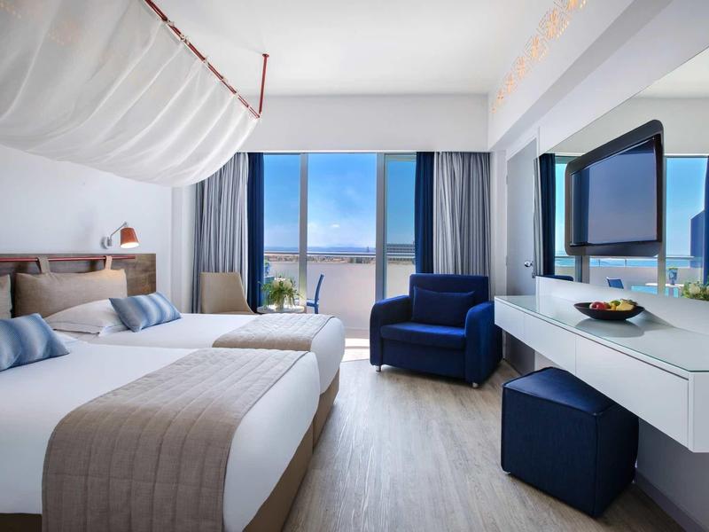 Modernes Hotelzimmer mit Doppelbett, blauem Sessel, großem Spiegel und Meerblick durch die Fenster.