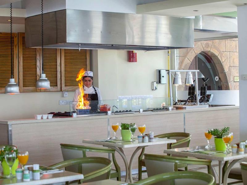 Moderner Gastraum mit Tischblumen, grünen Stühlen und offener Küche mit Koch und Flammen.