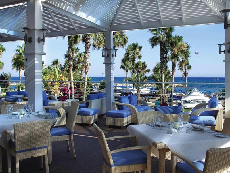 Terrassenrestaurant mit weißen Tischen, blauen Stühlen und Blick auf Palmen und Meer.