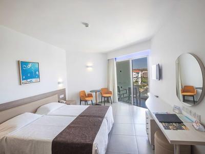 Světelný hotelový pokoj se dvěma postelemi, psacím stolem, židlemi a balkonem s výhledem ven.